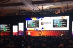 Large conference hall with stage and massive screens displaying mobile game graphics and logos, showcasing the latest advancements in mobile gaming technology and the gaming industry’s marketing events.