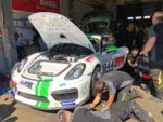 Mechanics working on a white Porsche race car with its hood open in a garage or pit area, demonstrating automotive repair and maintenance for high-performance vehicles.