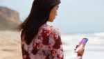 Profile view of a young Asian woman with long dark hair holding a smartphone and looking at the screen, standing on a beach with mountains in the background, illustrating mobile technology usage in scenic outdoor settings.