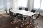 Long rectangular conference table with black rolling chairs arranged around it in a bright, modern office space with wood floors and large windows allowing natural light to stream in, showcasing a typical corporate meeting room setup.