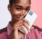 Close-up of a smiling young Black woman holding a smartphone and covering her mouth with her hand, demonstrating a casual and playful interaction with mobile technology against a pink background.