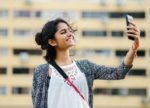 Smiling young Indian woman taking a selfie with her smartphone while outdoors in front of an apartment building, demonstrating casual mobile photography and social media sharing.